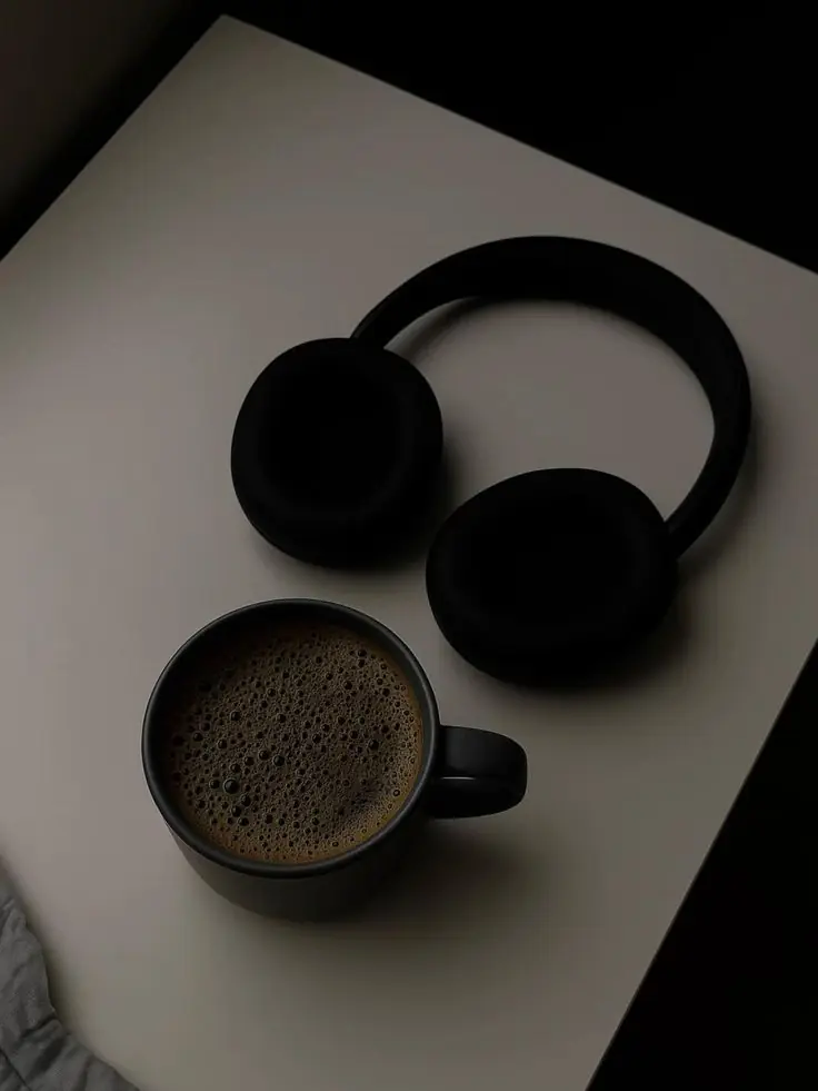 A black mug of coffee beside headphones on a pale table